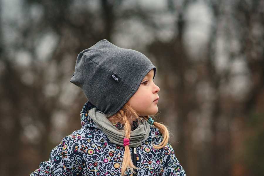 A little girl in a black and white patterned jacket with a gray cap with blond pigtails peeking out looks to the side